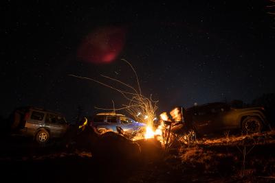 Two people sitting around a campfire, vehicles in the background